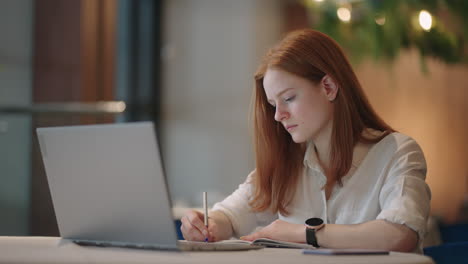 Cheerful-pretty-cute-nice-Red-haired-girl-girlfriend-having-been-employed-to-job-as-executive-smiling-toothily-sitting-at-desktop-with-laptop-noting-down-important-information